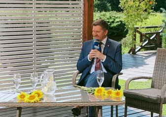 Członek Zarządu Województwa Lubelskiego Zdzisław Szwed w studiu telewizyjnym. Na stole dzbanek z wodą, dwa kieliszki na wodę oraz dekoracja ze słoneczników. W tle drewniany mostek oraz rośliny