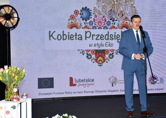 Na scenie stoi pan Zdzisław Szwed, Członek zarządu Województwa Lubelskiego . Zanim widać ekran z napisem "Kobieta Przedsiębiorcza w stylu Eko" na tle grafiki serca z kwiatów w stylu folkowym