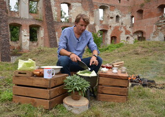 na zdjęciu w tle ruiny zamku w Krupem, na pierwszym planie Karol Okrasa siekający zioła, przed nim skrzynki drewniane pełniące role stolików, na nich zioła oraz produkty niezbędne do gotowania 