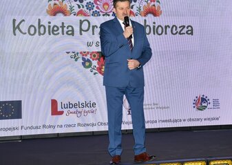 Pan Zdzisław Szwed, Członek Zarządu Województwa Lubelskiego stoi na scenie przy mikrofonie. Za nim widać biały ekran na którym widnieje grafika z sercem z kolorowych kwiatków w stylu folkowym z napisem „Kobieta Przedsiębiorcza w stylu Eko”