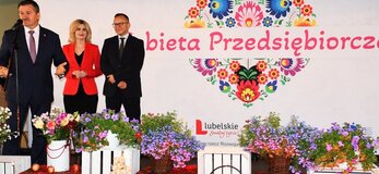 Trzy osoby stoją na scenie udekorowanej kwiatami. Od lewej strony Pan Zdzisław Szwed, Członek Zarządu Województwa Lubelskiego, w środku Pani Ewa Szałachwiej, Dyrektor Departamentu Rolnictwa i Rozwoju Obszarów Wiejskich i Pan Artur Soboń, Sekretarz Stanu w Ministerstwie Finansów. Osoby stoją na tle białej ścianki z dużym sercem z kwiatów w motywie folkowym i napisem przez jego środek "Kobieta Przedsiębiorcza"