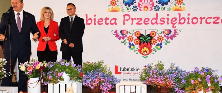 Trzy osoby stoją na scenie udekorowanej kwiatami. Od lewej strony Pan Zdzisław Szwed, Członek Zarządu Województwa Lubelskiego, w środku Pani Ewa Szałachwiej, Dyrektor Departamentu Rolnictwa i Rozwoju Obszarów Wiejskich i Pan Artur Soboń, Sekretarz Stanu w Ministerstwie Finansów. Osoby stoją na tle białej ścianki z dużym sercem z kwiatów w motywie folkowym i napisem przez jego środek "Kobieta Przedsiębiorcza" 