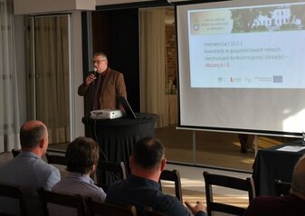 Mężczyzna ubrany elegancko, mówi do osób siedzących na krzesłach. Mężczyzna stoi obok ekranu na którym wyświetla się prezentacja