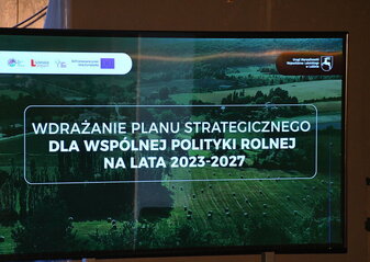 Grafika informująca o konferencji „Wdrażanie Planu Strategicznego dla Wspólnej Polityki Rolnej na lata 2023 – 2027”. Napis widnieje na tle zdjęcia pola na którym leżą bele siana i użytków zielonych, w dali widać las. Wszystko jest w tonacji ciemnozielonej.