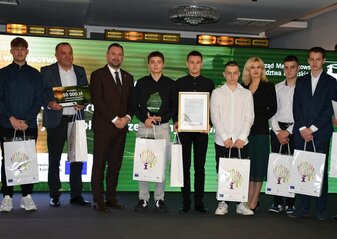 Na scenie, na tle kolorowej grafiki stoi grupa osób (kobieta i dziewięciu mężczyzn) ubranych elegancko, w dłoniach trzymają czek, statuetkę, dyplom i białą teczkę oraz białe torby prezentowe