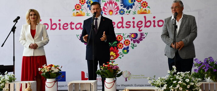 Trzy osoby stoją na scenie udekorowanej kwiatami. Jest to Pan Zdzisław Szwed, Członek Zarządu Województwa Lubelskiego, Pani Ewa Szałachwiej, Dyrektor Departamentu Rolnictwa i Rozwoju Obszarów Wiejskich oraz Pan Krzysztof Niewiadomski, Starosta Łęczyński