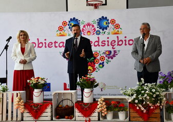 Trzy osoby stoją na scenie udekorowanej kwiatami. Jest to Pan Zdzisław Szwed, Członek Zarządu Województwa Lubelskiego, Pani Ewa Szałachwiej, Dyrektor Departamentu Rolnictwa i Rozwoju Obszarów Wiejskich oraz Pan Krzysztof Niewiadomski, Starosta Łęczyński