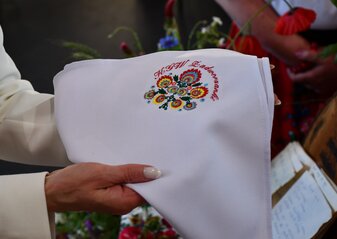 Wyszywany obrus prezentują kobiece dłonie. Biały obrus ma wyszyte kolorowe kwiaty w stylu ludowym i napis „KGW Zadworzanki”.