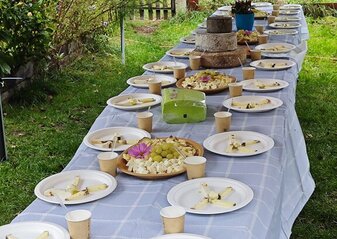Na dworze stoi długi stół nakryty białym obrusem. Na stole stoją talerzyki z porcjami degustacyjnymi sera wytwarzanego w gospodarstwie ekologicznym. 