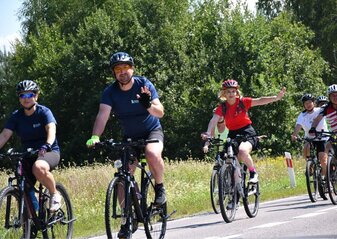 Grupa rowerzystów jedzie szosą. Mężczyzna i kobieta mają uniesione ręce w geście pozdrowienia 