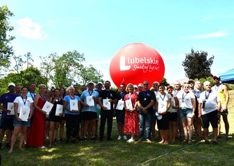 Grupa osób stoi na polanie. Są to uczestnicy spływu kajakowego. Za nimi widać balon promocyjny Lubelskie Smakuj Życie w kolorze czerwonym. Jest słonecznie, niebo ma błękitny kolor