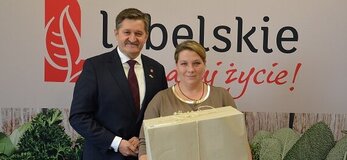 Na tle banera z napisem Lubelskie smakuj życie i grafiką zielonych ziół stoją dwie osoby. Po lewej stronie stoi Zdzisław Szwed, Członek Zarządu Województwa Lubelskiego. Obok niego stoi kobieta trzymająca duże pudełko owinięte w szary papier i przewiązane sznurkiem.