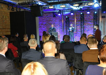 Marszałek województwa lubelskiego Jarosław Stawiarski stoi na scenie przy mównicy i mówi do uczestników konferencji „Wdrażanie Planu Strategicznego dla Wspólnej Polityki Rolnej na lata 2023 – 2027”. Obok niego stoi prowadzący konferencję.