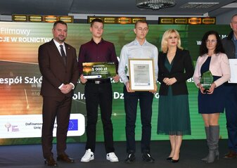 Na scenie, na tle kolorowej grafiki stoi grupa osób (dwie kobiety i czterech mężczyzn) ubranych elegancko, w dłoniach trzymają czek, statuetkę, dyplom i białą teczkę