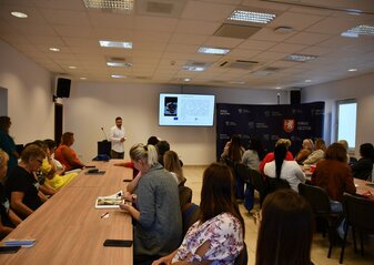 Kamil Kotarski na ekranie multimedialnym prezentuje przygotowaną prezentację, Uczestnicy konferencji siedzą przy stołach i słuchają