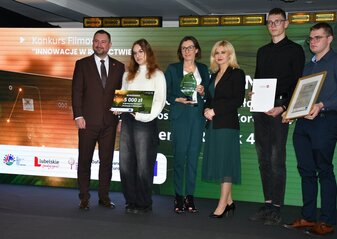 Na scenie, na tle kolorowej grafiki stoi grupa osób (trzy kobiety i trzech mężczyzn) ubranych elegancko, w dłoniach trzymają czek, statuetkę, dyplom i białą teczkę