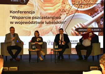 Na scenie na fotelach siedzą cztery osoby. Są to prelegenci oraz moderator, który prowadzi panel dyskusyjny podczas konferencji „Wsparcie pszczelarstwa w województwie lubelskim”.