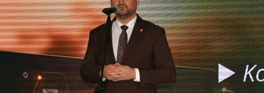 Mężczyzna ubrany elegancko stoi na scenie, przy mikrofonie na statywie. W tle wyświetla się kolorowa grafika z logotypami Unii Europejskiej, Planu Strategicznego dla Wspólnej Polityki Rolnej na lata 2023-2027, oraz napis Dofinansowane przez Unię Europejską.