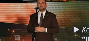 Mężczyzna ubrany elegancko stoi na scenie, przy mikrofonie na statywie. W tle wyświetla się kolorowa grafika z logotypami Unii Europejskiej, Planu Strategicznego dla Wspólnej Polityki Rolnej na lata 2023-2027, oraz napis Dofinansowane przez Unię Europejską.