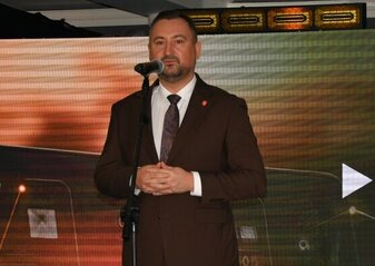 Mężczyzna ubrany elegancko stoi na scenie, przy mikrofonie na statywie. W tle wyświetla się kolorowa grafika z logotypami Unii Europejskiej, Planu Strategicznego dla Wspólnej Polityki Rolnej na lata 2023-2027, oraz napis Dofinansowane przez Unię Europejską.