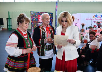 Trzy osoby stoją przy stole. Kobieta i mężczyzna ubrani w regionalne stroje ludowe to przedstawicie koła gospodyń wiejskich. Stoją obok pani Ewy Szałachwiej, Dyrektor Departamentu Rolnictwa i Rozwoju Obszarów Wiejskich, która czyta trzymaną w dłoniach zapisaną odręcznie kartkę papieru. Jest to stary przepis kulinarny. W tle widać scenę udekorowana kwiatami i białą ściankę z sercem z kwiatów w motywie folkowym i napisem przez środek serca „Kobieta Przedsiębiorcza”.
