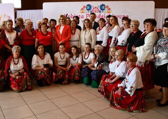 Duża grupa osób stoi na tle białej ścianki z sercem z kwiatów w motywie folkowym i napisem przez środek serca „Kobieta Przedsiębiorcza”. Są to przedstawicielki kół gospodyń wiejskich biorące udział w konkursie „Kobieta Przedsiębiorcza”. Panie ubrane są w regionalne stroje ludowe. Wśród nich jest pani Beata Mazurek, poseł do Parlamentu Europejskiego, pan Zdzisław Szwed, Członek Zarządu Województwa Lubelskiego i pani Ewa Szałachwiej, Dyrektor Departamentu Rolnictwa i Rozwoju Obszarów Wiejskich.