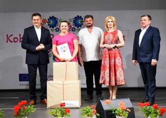 Reprezentantka Koła Gospodyń Wiejskich stoi na scenie z Panem Zdzisławem Szwedem, Członkiem Zarządu Województwa Lubelskiego i Panią Ewą Szałachwiej, Dyrektor Departamentu Rolnictwa i Rozwoju Obszarów Wiejskich. Obok stoją dwa duże pudełka z nagrodą która wygrała w konkursie