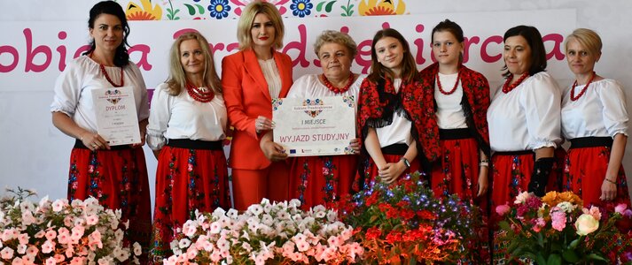 Osiem kobiet stoi na scenie udekorowanej świeżymi kwiatami. Są to przedstawicielki koła gospodyń wiejskich, których potrawa zwyciężyła w półfinale konkursu Kobieta Przedsiębiorcza. Panie ubrane są w regionalne stroje ludowe. Jedna z nich trzyma w dłoniach czek na wyjazd studyjny, który jest nagrodą w konkursie. Wśród pań jest pani Ewa Szałachwiej, Dyrektor Departamentu Rolnictwa i Rozwoju Obszarów Wiejskich. W tle widać scenę udekorowana kwiatami i białą ściankę z sercem z kwiatów w motywie folkowym i napisem przez środek serca „Kobieta Przedsiębiorcza”.