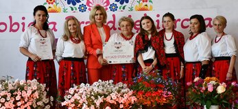 Osiem kobiet stoi na scenie udekorowanej świeżymi kwiatami. Są to przedstawicielki koła gospodyń wiejskich, których potrawa zwyciężyła w półfinale konkursu Kobieta Przedsiębiorcza. Panie ubrane są w regionalne stroje ludowe. Jedna z nich trzyma w dłoniach czek na wyjazd studyjny, który jest nagrodą w konkursie. Wśród pań jest pani Ewa Szałachwiej, Dyrektor Departamentu Rolnictwa i Rozwoju Obszarów Wiejskich. W tle widać scenę udekorowana kwiatami i białą ściankę z sercem z kwiatów w motywie folkowym i napisem przez środek serca „Kobieta Przedsiębiorcza”.