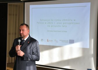 Mężczyzna ubrany elegancko, stoi na tle ekranu na którym wyświetla się napis Sytuacja na rynku chmielu w Polsce w 2025 roku oraz perspektywa na przyszłe lata, zestaw logotypów, znaczek Unii Europejskiej, napis Lubelskie Smakuj Życie, Znaczek KSOW+ oraz znaczek PS WPR 2023-2027 oraz napis Dofinansowane przez Unię Europejską