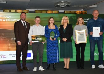 Na scenie, na tle kolorowej grafiki stoi grupa osób (trzy kobiety i trzech mężczyzn) ubranych elegancko, w dłoniach trzymają czek, statuetkę, dyplom i białą teczkę