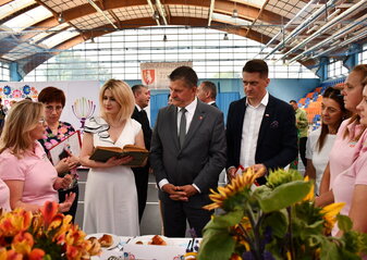 Komisja konkursowa ocenia produkty przygotowane przez koło gospodyń wiejskich. Wszyscy stoją przy stole, Pani Ewa Szałachwiej trzyma w rękach starą ksiażkę kucharską z której pochodzi przepis na przygotowaną przez panie z koła potrawę