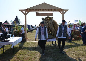 Mężczyźni w strojach ludowych niosący wieniec tradycyjny w tle brama drewniana z napisem Dożynki Wojewódzkie 2021