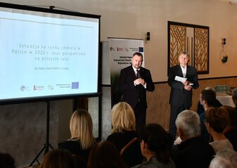 1. Dwóch mężczyzn ubranych elegancko, stoją przy ekranie na którym jest wyświetlany napis Sytuacja na rynku chmielu w Polsce w 2025 roku oraz perspektywa na przyszłe lata. Kilka siedzących osób