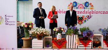 Trzy osoby, Pan Zdzisław Szwed, Członek Zarządu Województwa Lubelskiego, Pani Ewa Szałachwiej, Dyrektor Departamentu Rolnictwa i Rozwoju Obszarów Wiejskich w Urzędzie Marszałkowskim w Lublinie i Pan Henryk Karwan, Starosta Powiatu Tomaszowskiego stoją na scenie udekorowanej kwiatami. W tle widać białą ściankę z dużym sercem z kolorowych kwiatów w motywie folkowym i napisem "Kobieta Przedsiębiorcza"