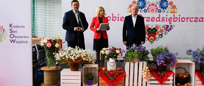 Trzy osoby, Pan Zdzisław Szwed, Członek Zarządu Województwa Lubelskiego, Pani Ewa Szałachwiej, Dyrektor Departamentu Rolnictwa i Rozwoju Obszarów Wiejskich w Urzędzie Marszałkowskim w Lublinie i Pan Henryk Karwan, Starosta Powiatu Tomaszowskiego stoją na scenie udekorowanej kwiatami. W tle widać białą ściankę z dużym sercem z kolorowych kwiatów w motywie folkowym i napisem "Kobieta Przedsiębiorcza"