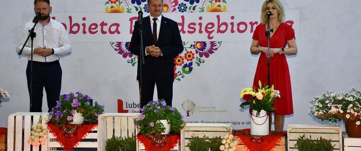 Trzy osoby stoją na scenie udekorowanej kwiatami. Jest to od lewej: Jacek Pożarowszczyk, Wicestarosta Powiatu Lubartowskiego, Zdzisław Szwed, Członek Zarządu Województwa Lubelskiego i Ewa Szałachwiej, Dyrektor Departamentu Rolnictwa i Rozwoju Obszarów Wiejskich. W tle widać białą ściankę z sercem z kwiatów w motywie folkowym i napisem przez środek serca :Kobieta Przedsiębiorcza"