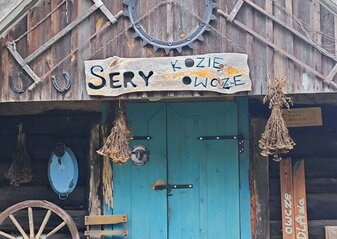 Drewniany domek w którym jest serowarnia. Stary drewniany domek z niebieskimi drzwiami z drewnianym szyldem „Sery owcze i kozie” ozdobiony jest podkowami, starymi narzędziami, bukietami suszonych kwiatów. Oparte jest o jego frontową ścianę koło od wozu, stoi niebieskie drewniane krzesło, wiklinowe kosze. Wszystko w sielskim folkowym stylu