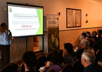 Grupa ludzi siedząca i słuchająca wykładu kobiety ubranej elegancko. Kobieta stoi przy ekranie na którym wyświetla się informacja o naborach wniosków w ramach Planu Strategicznego dla Wspólnej Polityki rolnej na lata 2023 – 2027