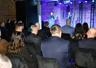 Marszałek województwa lubelskiego Jarosław Stawiarski stoi na scenie przy mównicy i mówi do uczestników konferencji „Wdrażanie Planu Strategicznego dla Wspólnej Polityki Rolnej na lata 2023 – 2027”. Obok niego stoi prowadzący konferencję.