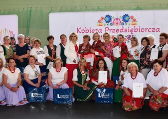 Duża grupa osób stoi ustawiona do wspólnego zdjęcia w Sali gimnastycznej. Są to kobiety i mężczyźni z kół gospodyń wiejskich i stowarzyszeń biorące udział w konkursie „Kobieta Przedsiębiorcza”. Niektórzy z nich trzymają w dłoniach torebki prezentowe. Wśród nich jest pani Anna Baluch, radna województwa lubelskiego i pani Ewa Szałachwiej, Dyrektor Departamentu Rolnictwa i Rozwoju Obszarów Wiejskich