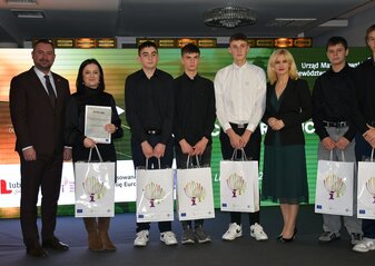 Na scenie, na tle kolorowej grafiki stoi grupa osób (dwie kobiety i sześciu mężczyzn) ubranych elegancko, w dłoniach trzymają białe torby prezentowe