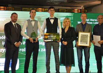 Na scenie, na tle kolorowej grafiki stoi grupa osób (kobieta i pięciu mężczyzn) ubranych elegancko, w dłoniach trzymają czek, statuetkę, dyplom i białą teczkę