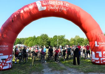 Grupa rowerzystów stoi pod czerwoną dmuchaną bramą na starcie rajdu. Wśród nich jest Pan Zdzisław Szwed, Członek Zarządu Województwa Lubelskiego oraz ksiądz, który odmówił modlitwę na bezpieczny przejazd uczestników