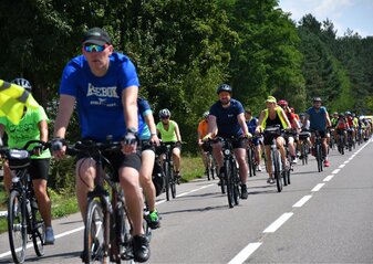 Duża grupa rowerzystów jedzie szosa. Za nimi widać las