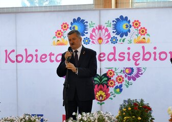 Pan Zdzisław Szwed, Członek Zarządu Województwa Lubelskiego stoi na scenie udekorowanej świeżymi kwiatami i mówi do mikrofonu. Za nim widać białą ściankę z sercem z kwiatów w motywie folkowym i napisem przez środek serca „Kobieta Przedsiębiorcza”.