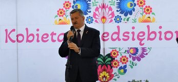 Pan Zdzisław Szwed, Członek Zarządu Województwa Lubelskiego stoi na scenie udekorowanej świeżymi kwiatami i mówi do mikrofonu. Za nim widać białą ściankę z sercem z kwiatów w motywie folkowym i napisem przez środek serca „Kobieta Przedsiębiorcza”.