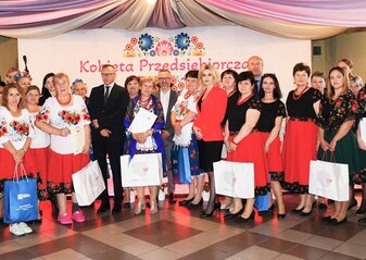 Grupa około 30 osób stoi na scenie udekorowanej kwiatami. Są to uczestniczki półfinału konkursu "Kobieta Przedsiębiorcza" powiatu świdnickiego. Wśród nich jest Pan Zdzisław Szwed, Członek Zarządu Województwa Lubelskiego, Pani Ewa Szałachwiej, Dyrektor Departamentu Rolnictwa i Rozwoju Obszarów Wiejskich i Pan Artur Soboń, Sekretarz Stanu w Ministerstwie Finansów. Osoby stoją na tle białej ścianki w dużym sercem w kwiatów w motywie folkowym i napisem przez jego środek "Kobieta Przedsiębiorcza"