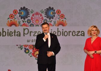 Pan Zdzisław Szwed, członek Zarządu Województwa Lubelskiego i Pani Ewa Szałachwiej, Dyrektor Departamentu Rolnictwa i Rozwoju Obszarów Wiejskich stoją obok siebie. Mężczyzna trzyma w dłoni mikrofon. Za nimi widać ekran z napisem Kobieta Przedsiębiorcza i grafikę serca z kwiatów w motywie folkowym