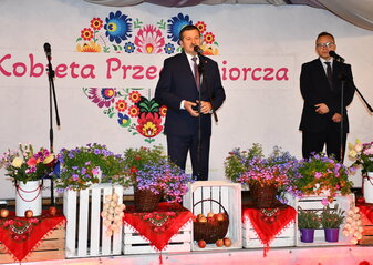 Dwóch mężczyzn stoi na scenie udekorowanej kwiatami. Od lewej strony Pan Zdzisław Szwed, Członek Zarządu Województwa Lubelskiego i Pan Artur Soboń, Sekretarz Stanu w Ministerstwie Finansów. Panowie stoją na tle białej ścianki z dużym sercem z kwiatów w motywie folkowym i napisem przez jego środek "Kobieta Przedsiębiorcza". 